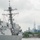 A U.S. Navy Arleigh Burke-class guided-missile destroyer is navigating waters near New York City, with the Manhattan skyline and One World Trade Center clearly visible in the background. Sailors in white dress uniforms are manning the rails of the ship, a traditional naval ceremony typically performed when entering or departing port. The scene appears to be part of Fleet Week or a similar naval event in New York Harbor.