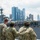 Several U.S. Navy personnel in digital camouflage uniforms (NWU Type III) are observed from behind, looking toward a large amphibious assault ship marked 'LHO 5' docked at a pier. A modern urban skyline with tall skyscrapers is visible in the background, suggesting a major U.S. port city. The scene appears to be during a port visit or fleet week event.