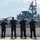Six uniformed law enforcement officers stand with raised hands facing a U.S. Navy vessel, on which a large military helicopter (appears to be an MH-53 or similar heavy-lift helicopter) is positioned on deck. An American flag is visible flying from the ship's superstructure, and a warning sign about jet blast and rotors is partially legible on the ship's hull. The scene appears to be at a port or pier during a public event or fleet week.