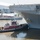 A red tugboat named 'Eric McAllister' is pushing against the hull of a large US Navy amphibious assault ship designated LHD-5 (USS Bataan). The scene appears to be at a busy waterfront pier, likely in New York City, with crowds of spectators visible on the dock and a British Airways aircraft visible in the background near what appears to be the Intrepid Sea, Air &amp; Space Museum. Naval personnel are visible on the ship's deck observing the docking operation.