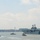 A large US Navy amphibious assault ship is navigating a harbor, accompanied by several tugboats. The scene appears to be in a major urban waterway, with a dense cityscape visible in the background. The overcast, hazy sky and calm water suggest mild weather conditions.