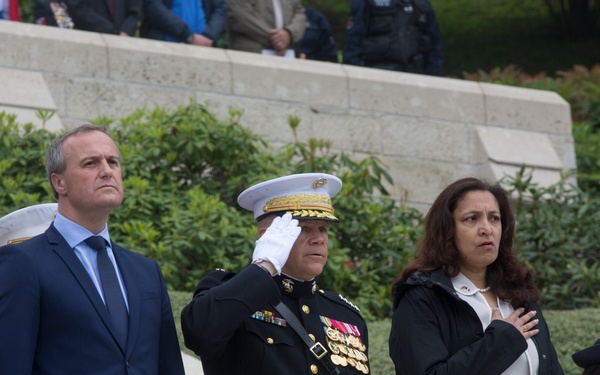 A formal military or commemorative ceremony is taking place, likely at a memorial or cemetery. A U.S. Marine Corps officer in dress blues is rendering a salute while civilians and other military personnel stand at attention. Several individuals are placing their hands over their hearts, suggesting a national anthem or moment of honor is being observed.