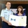 A United States Coast Guard officer in dress uniform and a woman in a blue dress are jointly holding an official certificate or commission document from the United States Coast Guard. The document features a blue flag with stars and the Presidential seal, suggesting a formal military ceremony. The setting appears to be an indoor venue with a large framed display in the background.