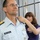 A U.S. Coast Guard officer named Michel is having insignia or a collar device pinned onto his uniform by a woman, likely during a promotion or change-of-command ceremony. The officer wears a light blue Coast Guard uniform with gold shoulder boards, a name tag reading 'MICHEL U.S. COAST GUARD,' service ribbons, and a warfare device. A second uniformed Coast Guard officer is partially visible in the background.
