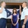 A formal military swearing-in or promotion ceremony involving two United States Coast Guard officers. Both officers have their right hands raised in an oath gesture, while a woman in a blue dress stands between them as a witness. The setting appears to be an official Coast Guard facility with flags and a flag display in the background.