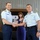 Two United States Coast Guard officers in light blue dress uniforms are shaking hands during what appears to be a formal military ceremony, likely a promotion or change of command. A woman in a blue dress stands between them, smiling. An American flag and a Coast Guard flag are visible in the background, along with a plaque or seal on the wall.