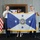 Three individuals — two uniformed U.S. Coast Guard officers and a civilian woman — are holding a U.S. Coast Guard Vice Commandant or senior flag with four blue stars and the Coast Guard seal, framed by gold fringe. The setting appears to be a formal military ceremony, likely a change of command or promotion event. A U.S. flag with gold fringe stands to the left, and a flag board with stars is visible in the background.