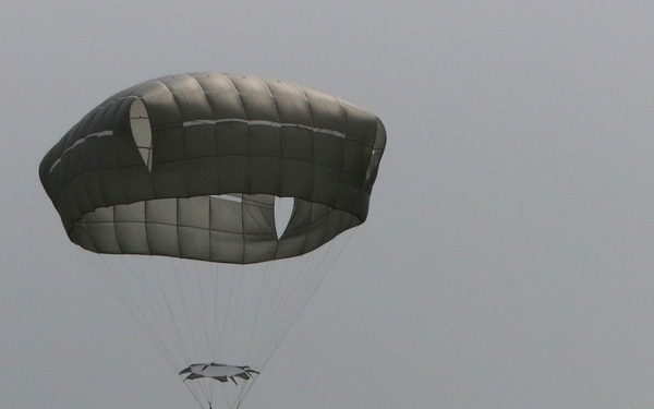 A military paratrooper descends under a large olive-drab/gray round parachute canopy in overcast, hazy conditions. A reserve parachute is also visible below the main canopy. In the background to the right, a military transport aircraft — consistent with a C-130 Hercules or similar turboprop — is visible flying away through the fog.