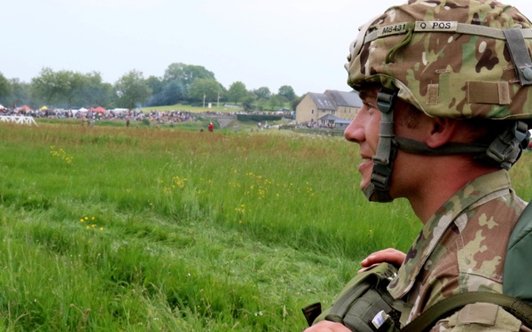 A U.S. Army soldier from the 82nd Airborne Division stands in a green field wearing full combat gear including a camouflage helmet and parachute pack. The soldier is facing left in profile view, observing a large crowd gathered in the background near what appears to be a commemorative or airborne demonstration event. The setting appears to be a European countryside, likely during a D-Day or airborne anniversary commemoration.