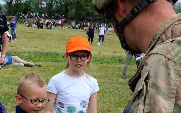 A soldier in camouflage uniform and helmet interacts with two young children at what appears to be an outdoor military demonstration or public event. A young boy wearing glasses and a blue vest is receiving or exchanging a small object with the soldier, while a girl in an orange cap and white shirt looks on. A large crowd of spectators is visible in the background on a grassy hillside.