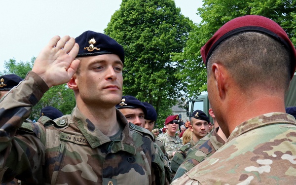 A military ceremony or greeting is taking place between soldiers from at least two different nations. A soldier in French Army camouflage uniform and dark navy beret is rendering a salute to a soldier in US Army MultiCam/OCP pattern uniform wearing a maroon/red beret. Multiple soldiers are visible in the background, suggesting a formal multinational military event or exercise.