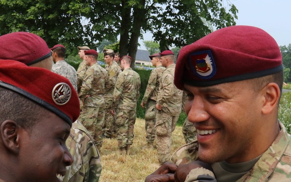 A military ceremony is taking place outdoors, likely a decoration or award pinning event between soldiers from two different nations. A soldier in French Army camouflage uniform with a red beret is pinning something onto a U.S. Army soldier wearing OCP (Operational Camouflage Pattern) uniform with a maroon beret. The U.S. soldier is smiling broadly, suggesting this is a positive recognition event, possibly a joint allied forces ceremony.