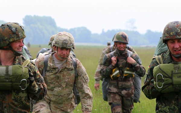 A group of soldiers from at least two different nations are walking across an open grassy field carrying parachute packs on their backs. The soldiers appear to have recently completed a parachute jump, as evidenced by the packed parachutes they are carrying. The mixed uniforms suggest this is a multinational military exercise or joint training operation.