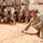 A military training session is taking place in an arid, desert environment. A soldier in MARPAT desert camouflage is kneeling on sandy ground, drawing or pointing to a diagram in the sand while addressing a group of soldiers in desert camouflage uniforms who are seated and observing attentively. The setting appears to be a forward operating base or training compound with buildings visible in the background.