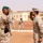 Two military officers from different nations are exchanging salutes in what appears to be a formal military ceremony in a desert environment. One officer wears a green beret with desert camouflage uniform, while the other wears a pixelated desert camouflage uniform consistent with U.S. Marine Corps MARPAT pattern. A formation of soldiers in the background, some carrying rifles, suggests this is an official military visit or ceremony.