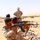 A military training exercise taking place in an arid desert environment. Two soldiers in desert camouflage uniforms are in a firing position aiming AK-pattern rifles, while a third soldier in MARPAT (Marine pattern) camouflage and boonie hat observes and supervises. The setting appears to be a forward operating base or training compound in a North African or Middle Eastern desert region.