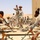 A military training exercise is taking place in a desert environment. A Western military instructor in distinctive camouflage uniform and boonie hat is demonstrating weapon handling to a group of local soldiers seated around a wooden platform with disassembled rifles. The setting appears to be an arid, sandy region with dunes visible in the background.