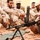 A U.S. Marine instructor is demonstrating the operation of a machine gun mounted on a bipod to a group of soldiers wearing desert camouflage uniforms. The setting appears to be an outdoor training environment with sandy, rocky ground near a building. This appears to be a military training or advisory session involving personnel from at least two different armed forces.