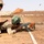 A military training exercise is taking place in an arid desert environment. One soldier in US Marine Corps desert MARPAT camouflage is instructing another soldier wearing a green helmet and desert camouflage uniform who is in a prone firing position. A spent cartridge casing is visibly ejecting from the AK-pattern rifle being fired.