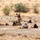 The image shows a military training exercise in an arid desert environment. One standing instructor in digital camouflage uniform appears to be directing or instructing multiple soldiers who are in prone firing positions on the ground. The trainees are wearing older-style steel helmets and lighter-colored uniforms, suggesting a multinational training scenario.