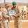 Two military officers — one in U.S. Marine Corps MARPAT desert camouflage with a patrol cap, and one in desert tri-color camouflage with a green beret — are exchanging a framed commemorative gift during what appears to be a formal military ceremony. The setting is an arid, desert environment with a formation of soldiers in the background wearing desert camouflage and red berets. The event appears to be a bilateral military cooperation or training exercise ceremony.