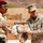 Two military personnel are exchanging a framed gift depicting camels in a desert setting. One individual wears a desert camouflage uniform with a red beret, consistent with a North African or Middle Eastern military force, while the other wears MARPAT (Marine Pattern) camouflage with a name tape partially reading 'SON', identifying him as a U.S. Marine. The exchange appears to be a ceremonial gift presentation accompanied by a handshake.