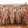 A group of approximately 18-20 U.S. Marines in desert MARPAT (digital camouflage) uniforms pose for a group photo in what appears to be a desert environment, likely North Africa or the Middle East. One service member on the right holds an American flag with gold fringe on a flagpole topped with an eagle finial. The setting includes a sandy terrain, palm trees, and a yellow building in the background.