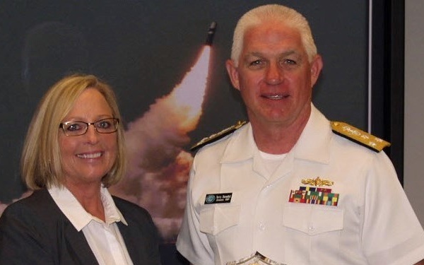 A naval officer in white dress uniform is presenting a crystal award to a civilian woman in a business jacket. The award appears to be a 'Director's Award' presented to 'Ms. Kimberley Payne' based on the visible engraving. They are posing together in what appears to be an official ceremony setting with a missile launch photograph displayed in the background.