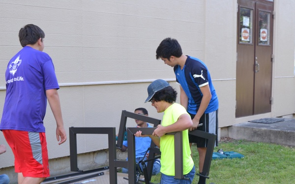 A group of youth and at least one adult are assembling dark metal furniture, likely a bench or table, on a grass lawn outside a building. The activity appears to be a community service or school project, with multiple participants working together on the assembly. Various metal frame components are laid out on the ground nearby.