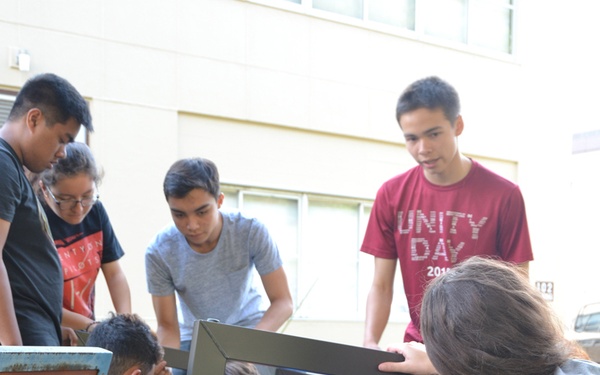 A group of approximately seven young people are gathered outdoors on a grass lawn, collaboratively assembling or repairing a piece of furniture, likely a bench or table with a metal frame. They appear to be working together as a team, with some kneeling and others standing to observe or assist. A building is visible in the background, and an older blue metal bench is visible to the left.