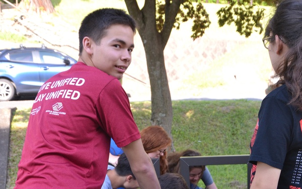 A group of young people are working together outdoors to assemble a piece of furniture, likely a picnic table or bench, on a grassy area. The boy in the foreground is wearing a red 'Boys &amp; Girls Clubs of America' t-shirt with 'Live Unified, Play Unified' text on the back. An instruction sheet is visible on the ground, and the group appears to be using power tools for the assembly.