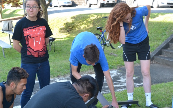 A group of young people, likely students, are assembled outdoors on a grassy area assembling or repairing what appears to be a park bench or picnic table. Several individuals are actively working on the metal frame structure on the ground while others observe. The setting appears to be a school or community park environment, suggesting a volunteer or service project.