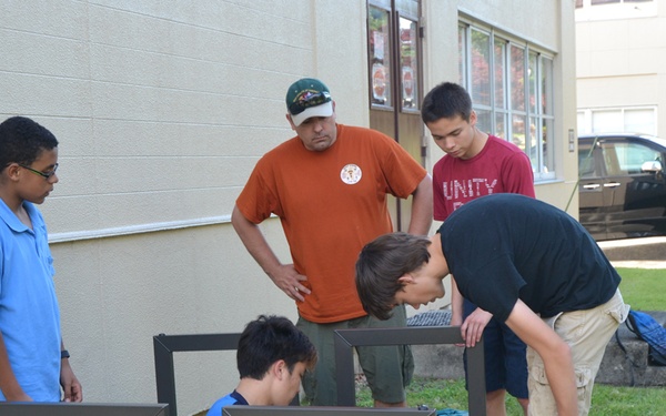 A group of four young teenagers and one adult male are assembled outdoors on a grass lawn beside a building, working together to assemble what appears to be metal furniture or equipment frames. One youth is seated on the ground using a power drill while others observe and assist. The scene suggests a supervised hands-on construction or community service activity.