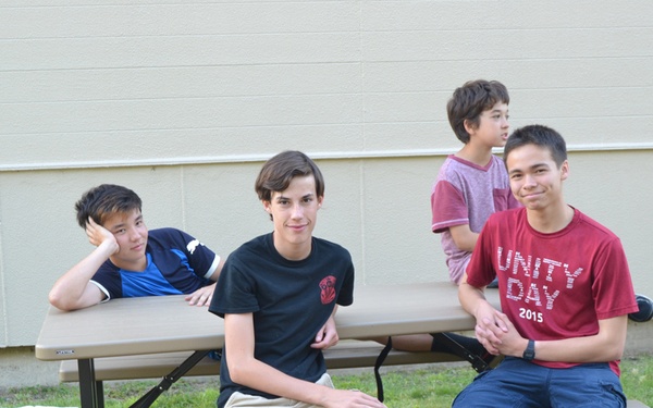 Four young males are gathered around a plastic picnic table on a grass lawn beside a building exterior wall. Two are seated on the bench facing the camera, one is leaning on the tabletop, and one stands in the background. The setting appears to be a school or community outdoor area.