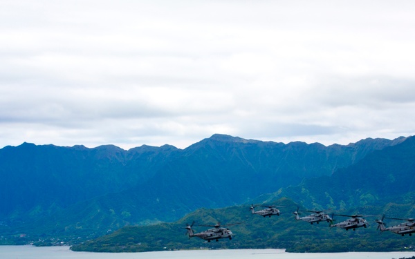 A formation of military helicopters flies over a coastal bay surrounded by lush green mountains and a narrow peninsula. The scene appears to be in Hawaii, likely near Kaneohe Bay on the island of Oahu, based on the distinctive mountain ridgelines and coastal geography. The helicopters are flying in a loose echelon formation at low altitude over the water.