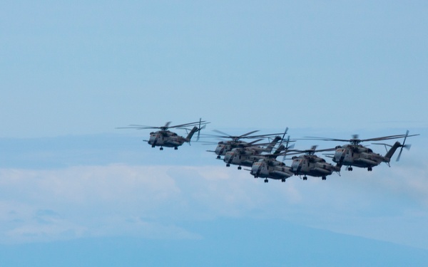 A formation of five large military helicopters flying in close proximity over an open body of water. The helicopters appear to be heavy-lift variants, likely CH-53 Sea Stallions or similar models, painted in a tan/desert camouflage scheme. A distant landmass or mountain range is visible on the horizon beneath a hazy blue sky.