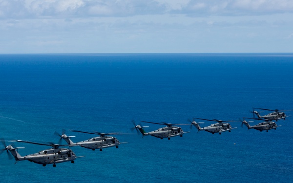A formation of seven large military helicopters flying in a diagonal echelon formation over open ocean. The helicopters appear to be CH-53 Super Stallion or similar heavy-lift variants, painted in gray military livery. The image was taken from an aerial vantage point, likely from another aircraft, with clear blue ocean and partial cloud cover visible.