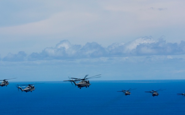 A formation of six military helicopters flying in loose echelon formation over open ocean. The helicopters appear to be large, heavy-lift military variants, likely CH-53 Sea Stallions or similar models, flying at low altitude above deep blue water. The sky is partly cloudy with cumulus clouds visible on the horizon.