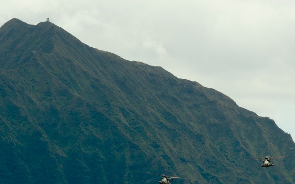 A formation of five military helicopters flies in a loose echelon formation against a backdrop of steep, heavily vegetated mountain terrain under overcast skies. The helicopters appear to be twin-rotor tandem-rotor type aircraft, consistent with CH-46 Sea Knight or MH-60 variants, flying at low-to-medium altitude. The lush green ridgelines and dramatic peak suggest a tropical or subtropical island environment, possibly Hawaii.