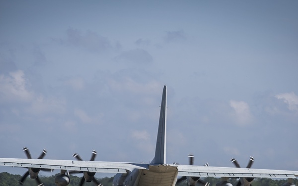 A large military turboprop aircraft, consistent with a C-130 Hercules variant, is photographed from the rear on a runway or taxiway. All four propeller engines are spinning, indicated by motion blur on the propeller blades. The aircraft appears to be preparing for takeoff or taxiing, set against a backdrop of green treeline and partly cloudy sky.