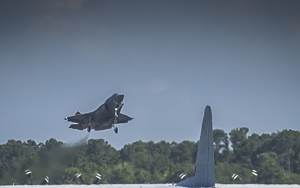 The image shows a military airfield scene with two distinct aircraft visible. A large four-engine turboprop transport aircraft, consistent with a C-130 Hercules, is on or near the runway in the foreground, while a smaller delta-wing jet fighter is captured airborne in the background, appearing to be taking off or performing a low pass. The scene is set against a backdrop of dense green treeline under an overcast sky.