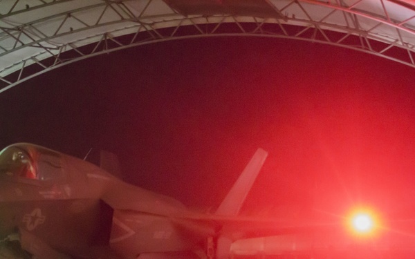 A military stealth fighter jet, consistent with an F-35 Lightning II, is positioned inside or under an arched hangar structure at night. The scene is dramatically lit by a bright red/orange light source on the right side, along with a green navigation light visible on the aircraft's left wing. At least one ground crew member is visible to the right of the aircraft.