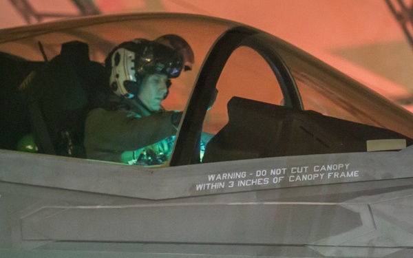A pilot in full flight gear, including helmet and oxygen mask, is seated in the cockpit of what appears to be an F-35 Lightning II stealth fighter jet. The aircraft is photographed in low-light conditions, likely inside a hangar, with orange ambient lighting visible in the background. Several safety and rescue markings are clearly visible on the fuselage.