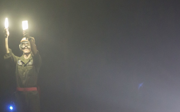 A person in military or aviation ground crew attire is standing on what appears to be a tarmac or airfield at night, holding two illuminated wands or light paddles raised above their head. The scene is shrouded in fog or haze, creating a dramatic, low-visibility atmosphere. The individual appears to be performing aircraft marshalling signals.