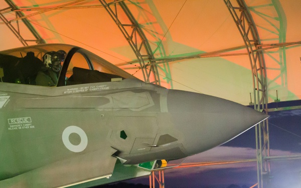 A fifth-generation stealth fighter jet, identifiable as an F-35, is shown inside a military shelter or hangar at night. The aircraft bears a roundel marking consistent with the Royal Air Force or a NATO ally, and ground crew personnel are visible performing pre-flight or post-flight checks. Coordinate and elevation data are stenciled on the hangar structure in the upper right corner.