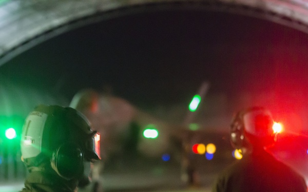 A nighttime military flight line scene showing two personnel wearing helmets and hearing protection standing under a large arched hangar structure. An aircraft is visible in the background amid colored navigation or warning lights. The scene is illuminated by green and red lights, creating a dramatic low-light operational atmosphere.