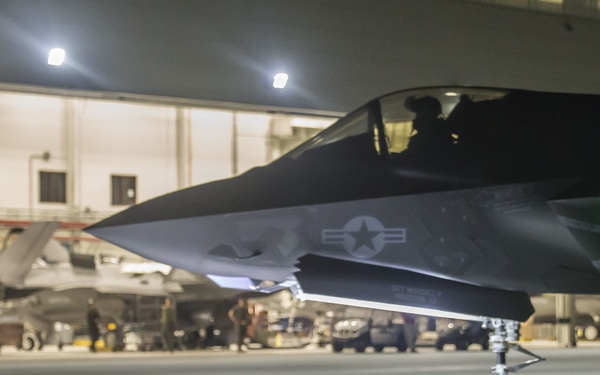 A U.S. military F-35 stealth fighter jet is photographed at night on a flight line apron, taxiing or parked near an illuminated hangar. The aircraft displays a U.S. Air Force/Navy roundel insignia on its fuselage and has its cockpit occupied by a pilot. Additional aircraft and ground personnel are visible in the background near the hangar.