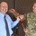 A U.S. Army soldier in OCP (Operational Camouflage Pattern) uniform is presenting a red lanyard to a civilian man in a blue dress shirt and tie. Both individuals appear to be in good spirits, with the civilian laughing. The setting appears to be outside a building with brown doors.