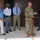 A military officer in Army OCP (Operational Camouflage Pattern) uniform is speaking at a small podium/lectern to a group of four civilians in business casual attire. The setting appears to be an outdoor covered area adjacent to a brick building. The gathering has the appearance of an official briefing, ceremony, or community engagement event.
