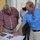Three older men are gathered in a corridor of what appears to be an institutional building, reviewing documents spread on a surface. The man in a blue shirt is pointing at what appears to be blueprints or plans, alongside a CD/DVD disc and a printed aerial photograph. The setting appears to be a facility walkway with cinder block walls painted blue and white.